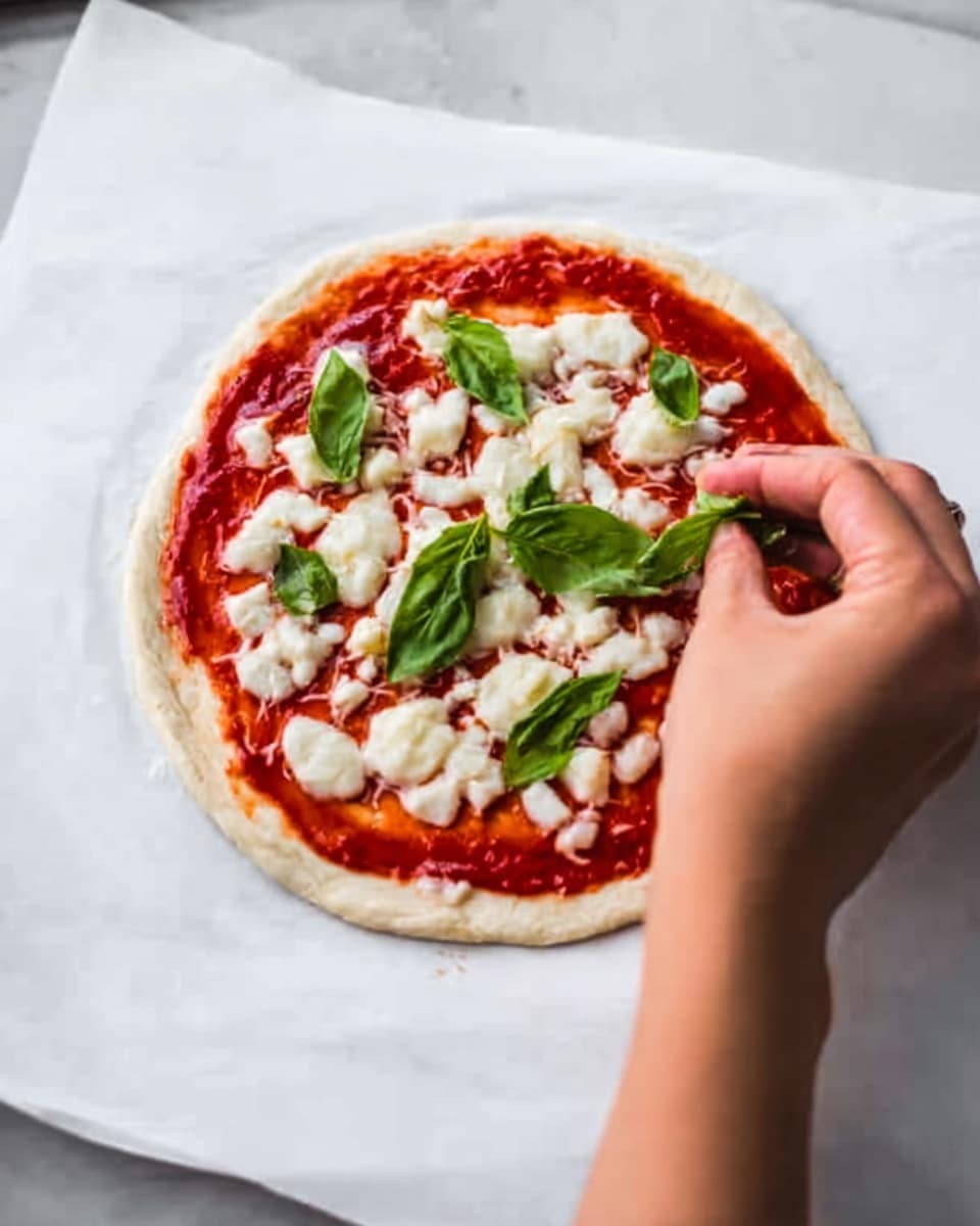 A small round pizza dough sits flat on white baking paper placed on a white marbled surface. The dough is covered evenly with bright red tomato sauce in the middle layer. On top of the sauce, there are small chunks of white melted cheese spread across the pizza. A woman's hand is placing fresh green basil leaves on the top, adding a fresh touch to the pizza. Photo taken with an iphone --ar 4:5 --v 7