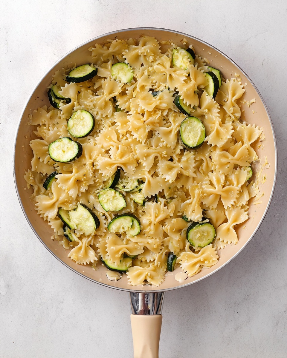 A beige pan with a long handle is filled with light yellow bowtie pasta mixed with small pieces of green zucchini slices. The pasta is cooked and slightly glossy, scattered evenly across the pan, with the zucchini slices showing soft green skin and pale inner flesh. The pan sits on a white marbled surface, creating a clean background that highlights the simple colors and textures of the dish. Photo taken with an iphone --ar 4:5 --v 7