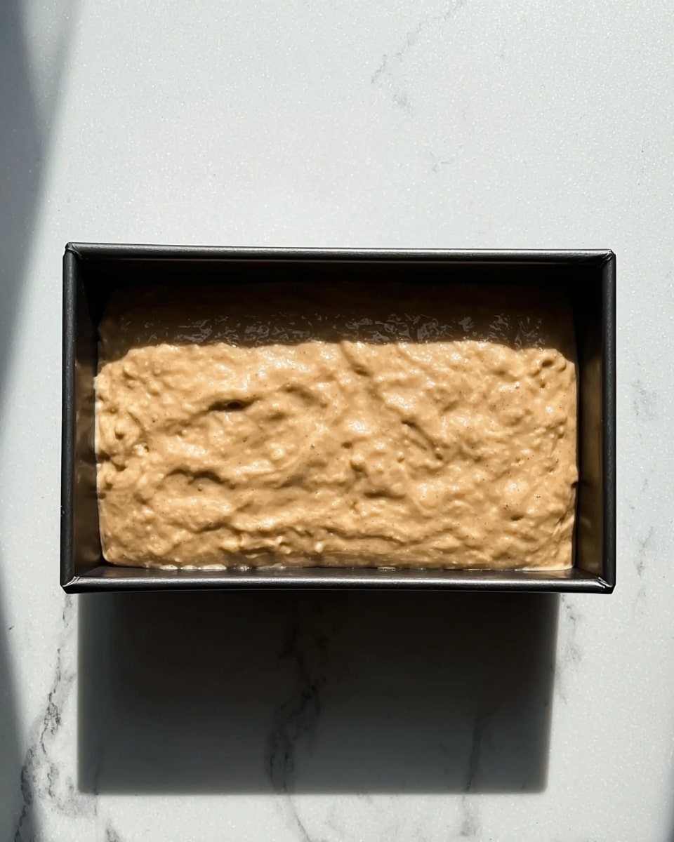 The image shows a rectangular black baking pan placed on a white marbled surface. Inside the pan is an uneven layer of thick light brown batter with a slightly lumpy texture spread across the entire surface. The batter has some small air pockets and subtle shiny spots reflecting light. There are soft shadows around the pan, indicating natural light from above. Photo taken with an iphone --ar 4:5 --v 7