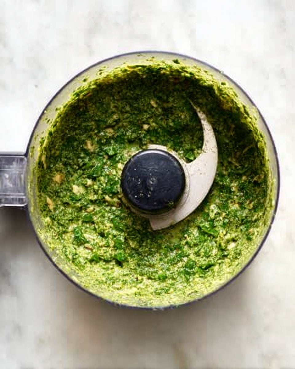 A clear food processor bowl filled with a thick, green pesto sauce that has a slightly coarse texture with small bits of herbs and nuts visible. The blade sits in the center, partially covered by the pesto. The bowl rests on a white marbled surface. Photo taken with an iphone --ar 4:5 --v 7
