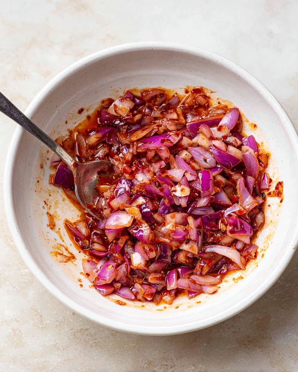 A white bowl filled with chopped red onions mixed with a reddish sauce, giving the onions a glossy, slightly sticky texture. There is a silver spoon resting inside the bowl on the left side, partially covered by the mixture. The bowl sits on a white marbled surface, and the sauce clings to the sides of the bowl, showing a thick consistency. photo taken with an iphone --ar 4:5 --v 7