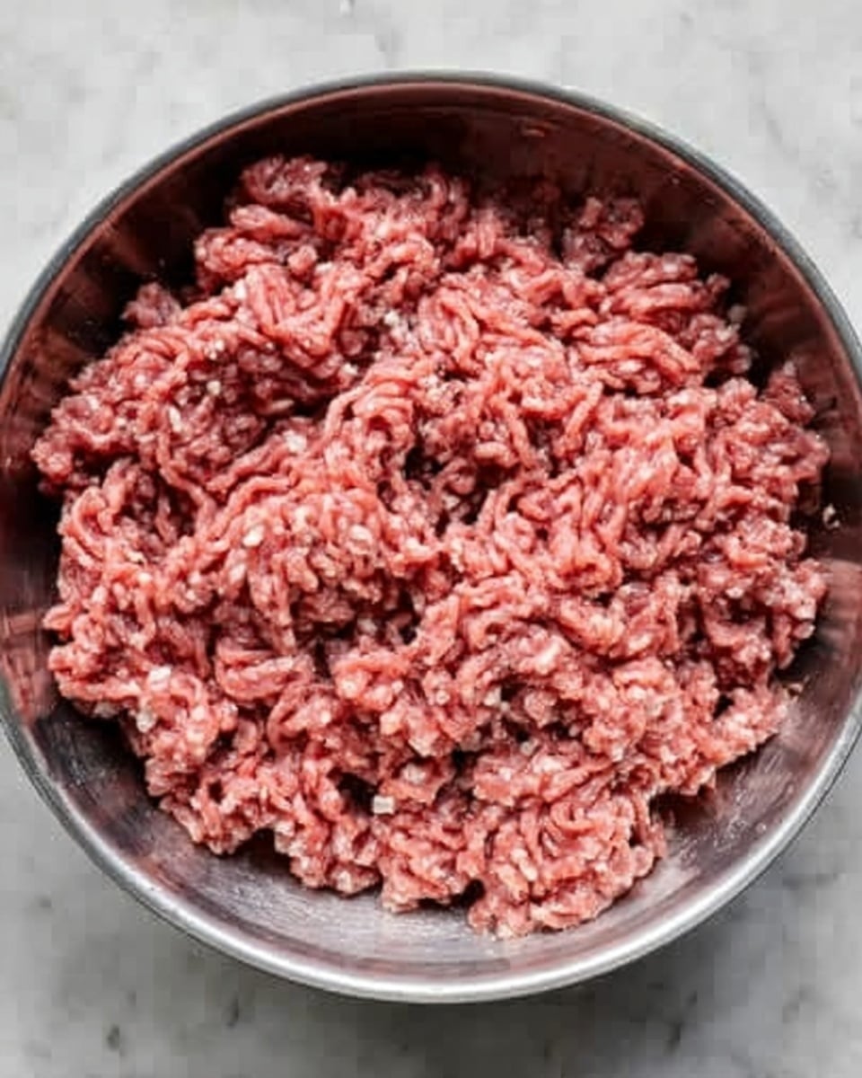 A close-up view of a silver pan filled with one layer of raw ground meat with a pinkish-red color and soft, crumbly texture evenly spread inside. The pan is placed on a white marbled surface, showing slight shine on the metal and the meat pieces with small white fat bits mixed in photo taken with an iphone --ar 4:5 --v 7