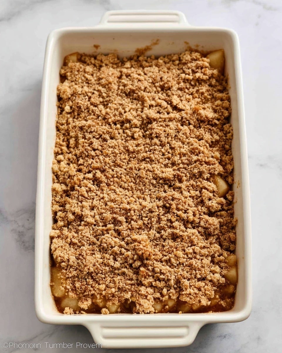 A rectangular white ceramic baking dish is filled with a dessert that has two visible layers. The bottom layer is a chunky mixture of soft, light brown pieces, likely fruit, mostly hidden beneath a thick, crumbly topping that covers the dish evenly. The topping is a medium brown color, appearing coarse with a grainy texture, suggesting a crunchy, streusel-like finish. The dish sits on a white marbled surface. Photo taken with an iphone --ar 4:5 --v 7