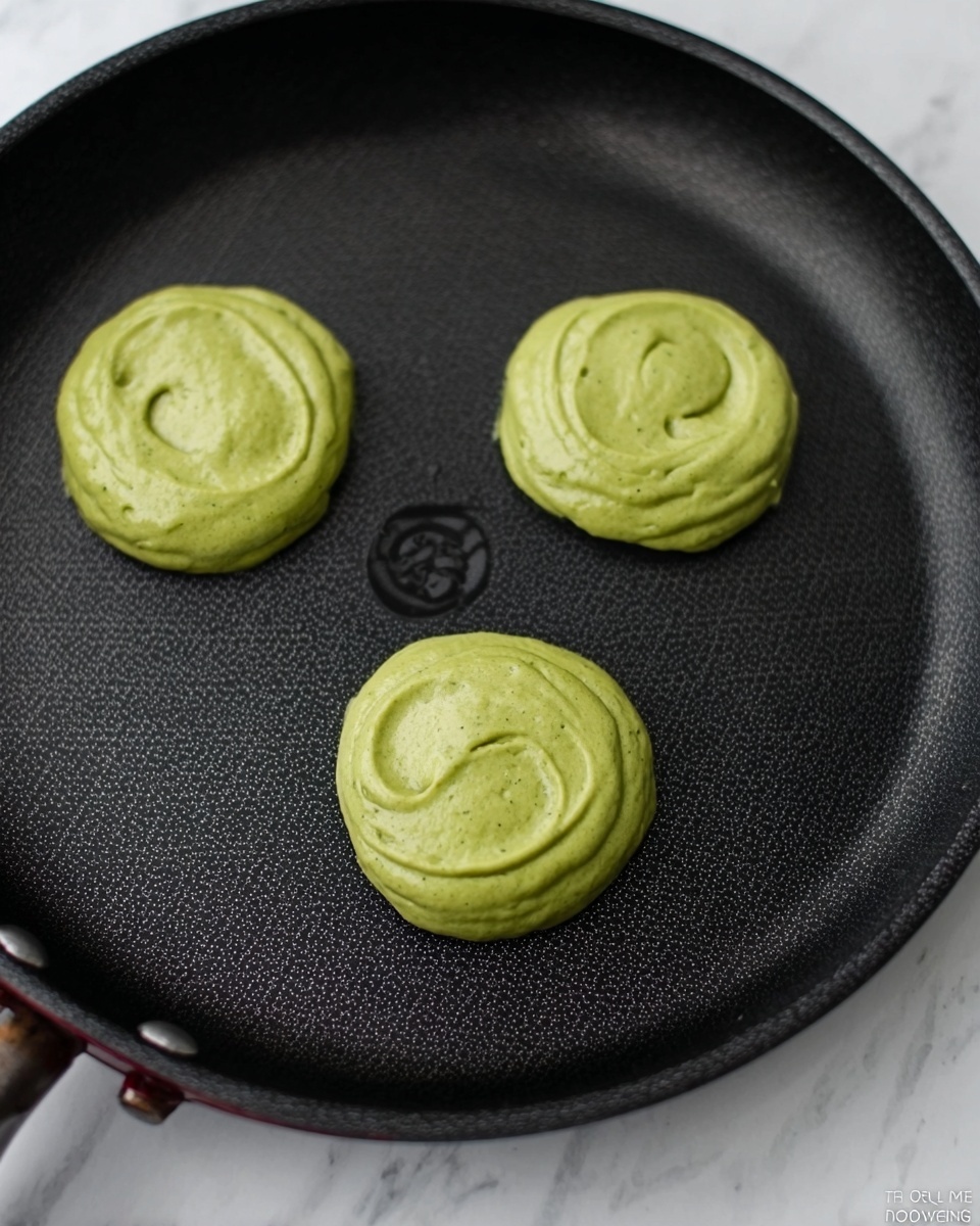 The image shows three small, round green batter dollops cooking on a black non-stick pan. Each dollop has a thick, smooth texture with slight swirls on top, and they are evenly spaced apart. The pan has a subtle pattern inside and a matte black rim. The background is a white marbled surface. photo taken with an iphone --ar 4:5 --v 7
