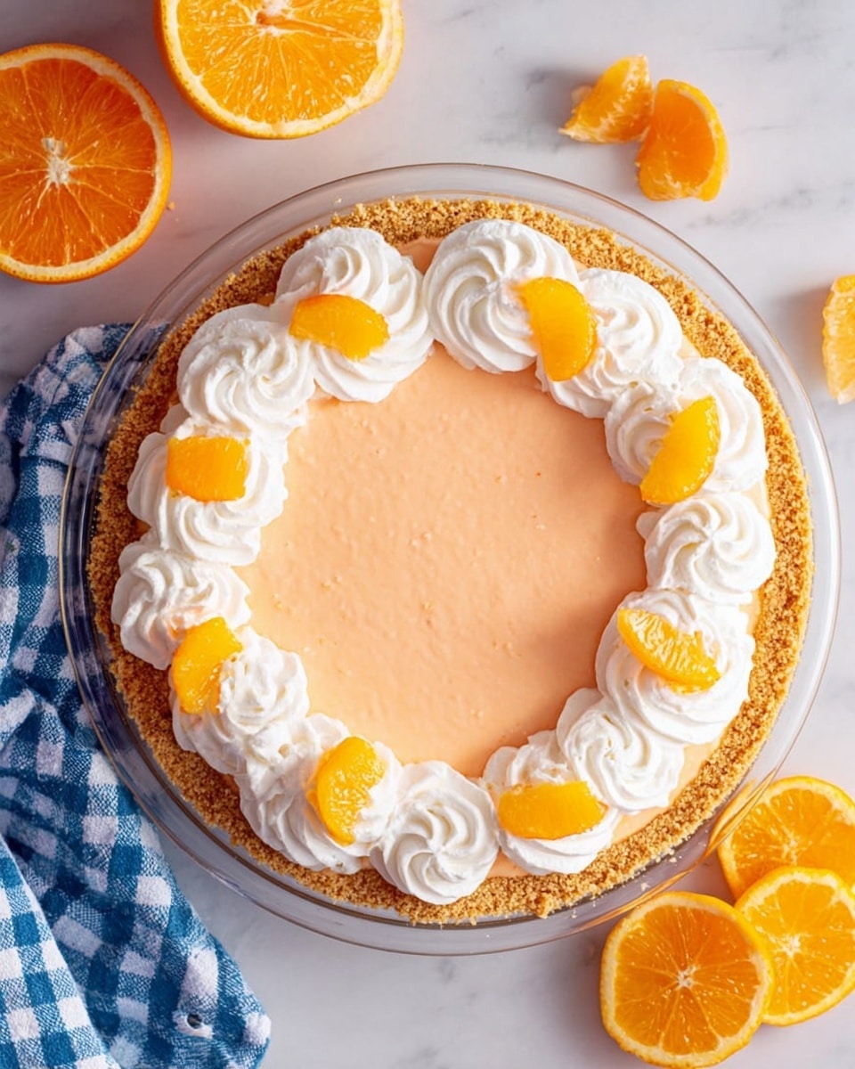 A round pie with three layers is shown from above on a white marbled surface. The bottom layer is a golden-brown crumb crust, the middle layer is smooth and light orange, and the top layer has white whipped cream swirls placed evenly around the edge. Small, bright orange orange segments sit on top of some whipped cream swirls, adding color contrast. An orange cut in half and some orange slices are placed near the pie with a blue and white checkered cloth partly visible to the left. photo taken with an iphone --ar 4:5 --v 7