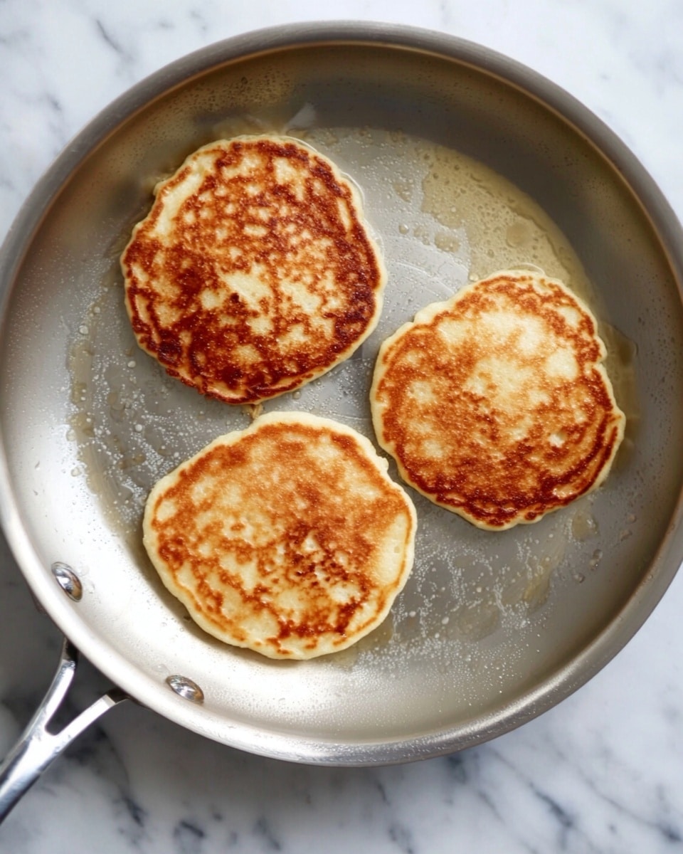 The image shows three golden brown pancakes cooking in a silver frying pan placed on a white marbled surface. The pancakes are round with slightly uneven edges and have small bubbles and darker spots on their surfaces, indicating they are being cooked evenly. The pan has a smooth, slightly shiny texture with some oil reflections. Photo taken with an iphone --ar 4:5 --v 7