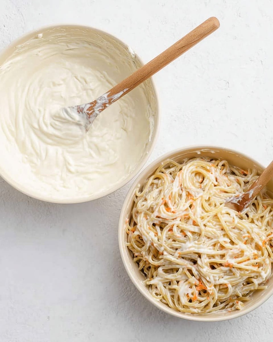 Two round white bowls are shown on a white marbled surface; the left bowl contains a thick, creamy white sauce with a wooden spoon resting inside, coated in the sauce, while the right bowl holds a mix of noodles coated in the same creamy sauce with visible small orange specks, and a wooden spoon stirring the noodles. photo taken with an iphone --ar 4:5 --v 7