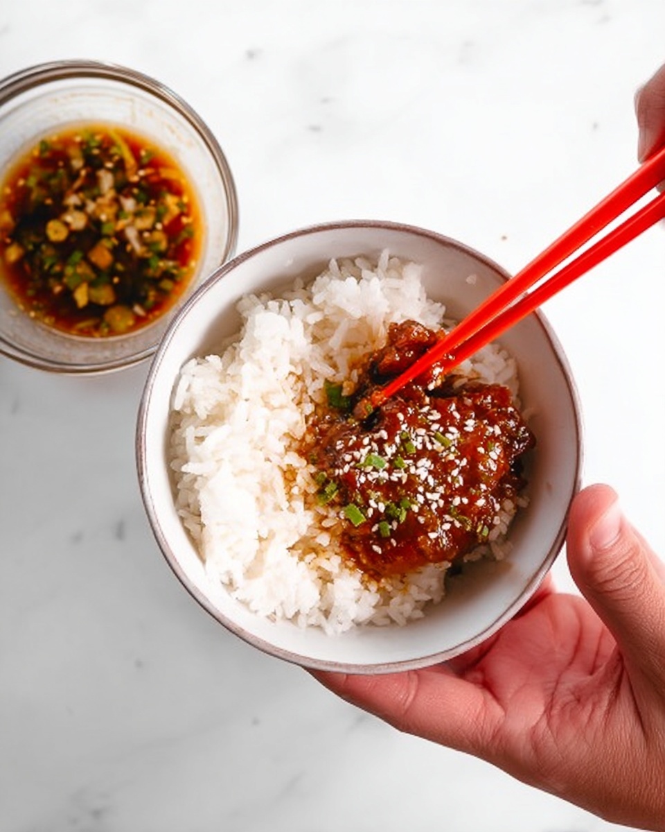 A small white bowl filled with white rice, topped with a shiny brown sauce-covered piece of food sprinkled with white sesame seeds and small green bits. A woman's hand holds the bowl from the side, while another woman's hand uses bright red chopsticks to pick up the piece of food. In the background, there is a small clear bowl with dipping sauce that has sesame seeds and chopped green chili peppers. The scene is set on a white marbled surface. photo taken with an iphone --ar 4:5 --v 7