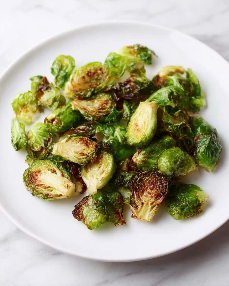 A white plate holds a single layer of roasted Brussels sprouts, each piece showing a mix of vibrant green and browned crispy edges. The leaves are curled and slightly charred, giving a rough texture on top with a fresh, soft green inside. The plate sits on a white marbled surface, and the lighting highlights the contrast between the crisp, golden brown parts and the fresh green leaves, making the dish look simple and appetizing. Photo taken with an iphone --ar 4:5 --v 7