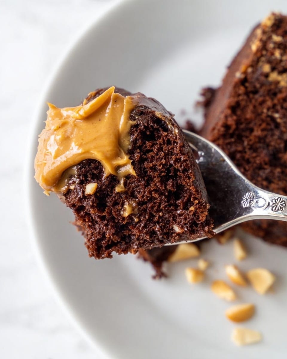 A close-up image of a moist, dark brown chocolate cake piece held on a spoon, with smooth peanut butter spread on top. The cake looks soft with a light holey texture, and the peanut butter has a creamy, slightly shiny look that spreads unevenly on the cake edges. The spoon is shiny silver with a floral pattern on the handle, resting on a white plate. Small bits of light beige chopped nuts are scattered on the plate near the spoon. The background is a white marbled surface. photo taken with an iphone --ar 4:5 --v 7