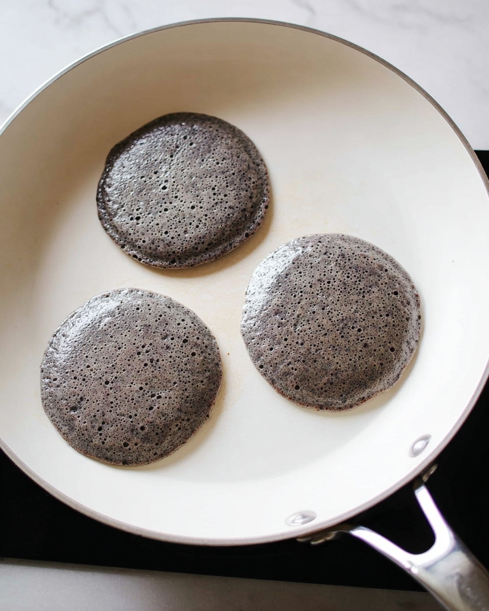 Three black and gray speckled batter pancakes cook in a white pan on a black stove. Each pancake is round and thick with small bubbles on the surface. The pancakes are evenly spaced in the pan and the glossy texture shows heat cooking them. The pan handle shines with a metallic finish and the setup sits on a clean white marbled surface photo taken with an iphone --ar 4:5 --v 7