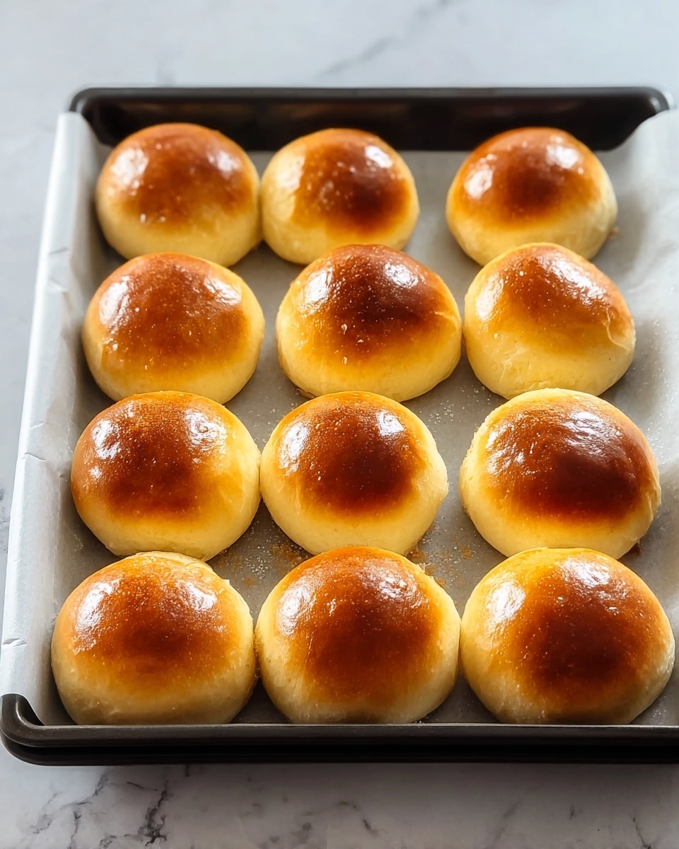 This image shows a black baking tray lined with white parchment paper, holding fifteen small round golden brown buns arranged in three rows. Each bun has a smooth, shiny top with a rich warm color gradient from dark golden brown at the center to lighter golden at the edges. The buns have a soft, smooth texture, and the light reflects off the glossy, slightly crisp surface. The tray sits on a white marbled surface with soft grey veins. Photo taken with an iphone --ar 4:5 --v 7