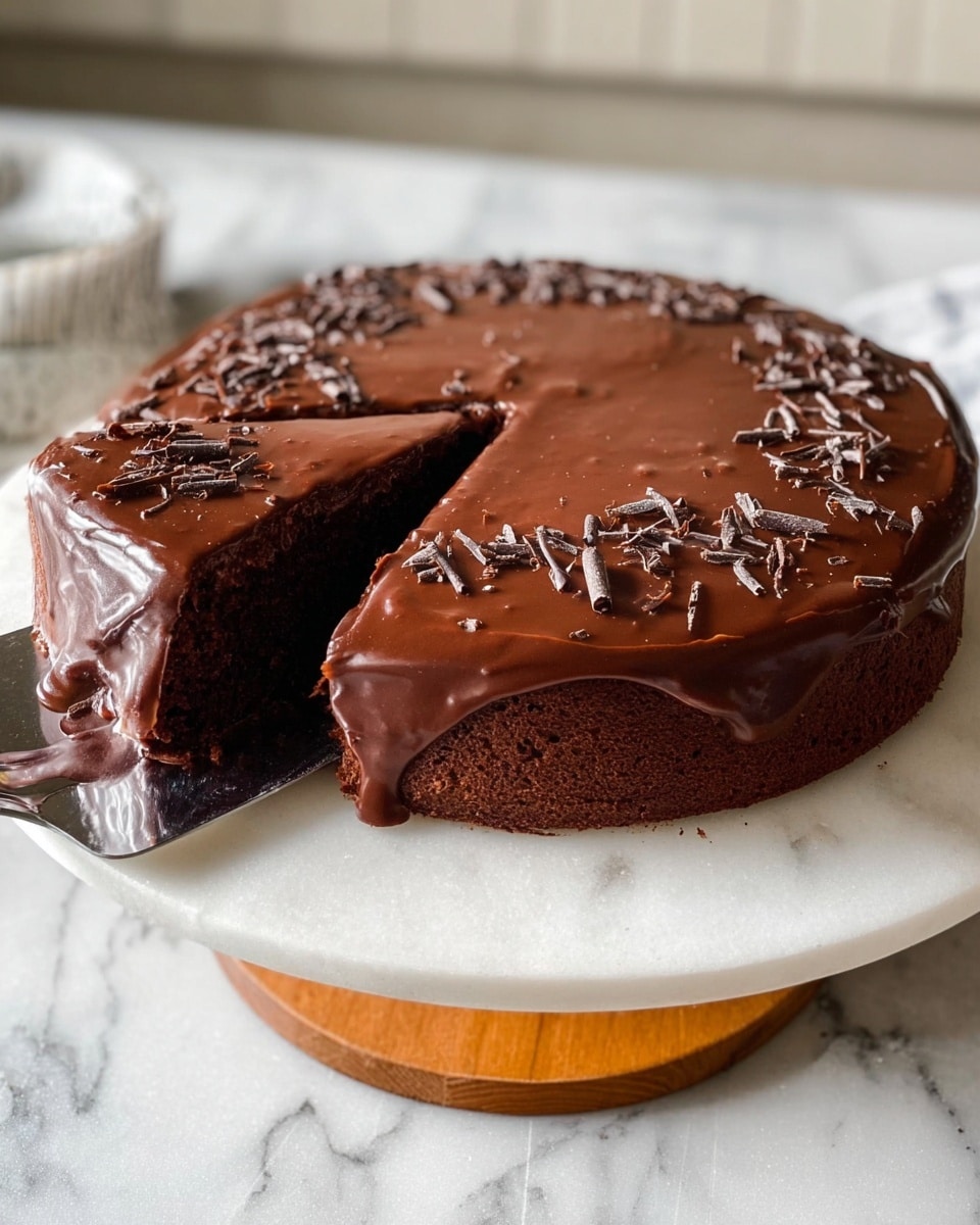 A round chocolate cake with one thick dark brown cake layer is covered by a smooth, shiny layer of rich dark chocolate glaze that spreads evenly on top and drips slightly on the sides. The top is decorated with small dark chocolate curls scattered mostly around the edges. A slice is lifted on a silver spatula revealing the moist inside and the glossy glaze on top. The cake sits on a white marble cake stand with a wooden base, placed on a white marble surface with soft light coming from the side. Photo taken with an iphone --ar 4:5 --v 7