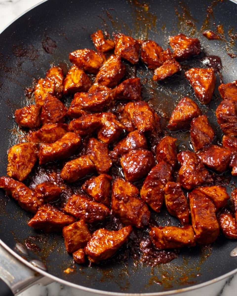 The image shows a black frying pan filled with many small shiny pieces of cooked meat, each covered in a thick, dark reddish-brown sauce. The meat pieces have a glossy texture with slight charring spots, and they are spread mostly in the center of the pan, with a few scattered around the edges. The pan's surface has a slightly rough texture with some oil and small burnt bits visible. The background beneath the pan is a white marbled surface. photo taken with an iphone --ar 4:5 --v 7