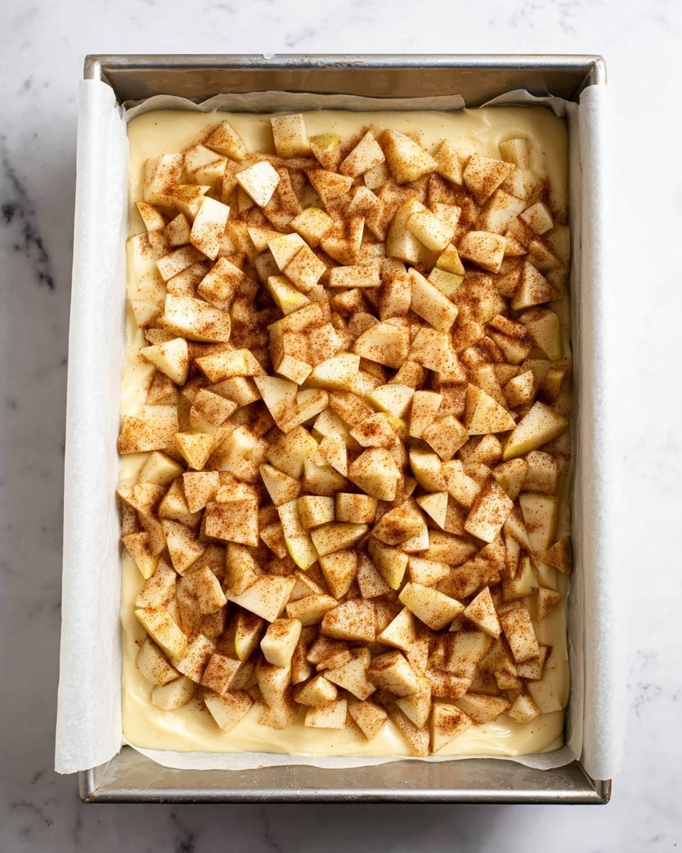 A rectangular metal baking pan lined with white parchment paper holds a layered dish. The bottom layer is a smooth, creamy batter in a pale yellow color, spread evenly across the pan. On top, there is a thick layer of small, chopped apple pieces in varying shapes, lightly coated with cinnamon, giving them a warm brown speckled look. The apples are spread evenly but with some gaps showing the batter beneath. The pan sits on a white marbled surface. photo taken with an iphone --ar 4:5 --v 7