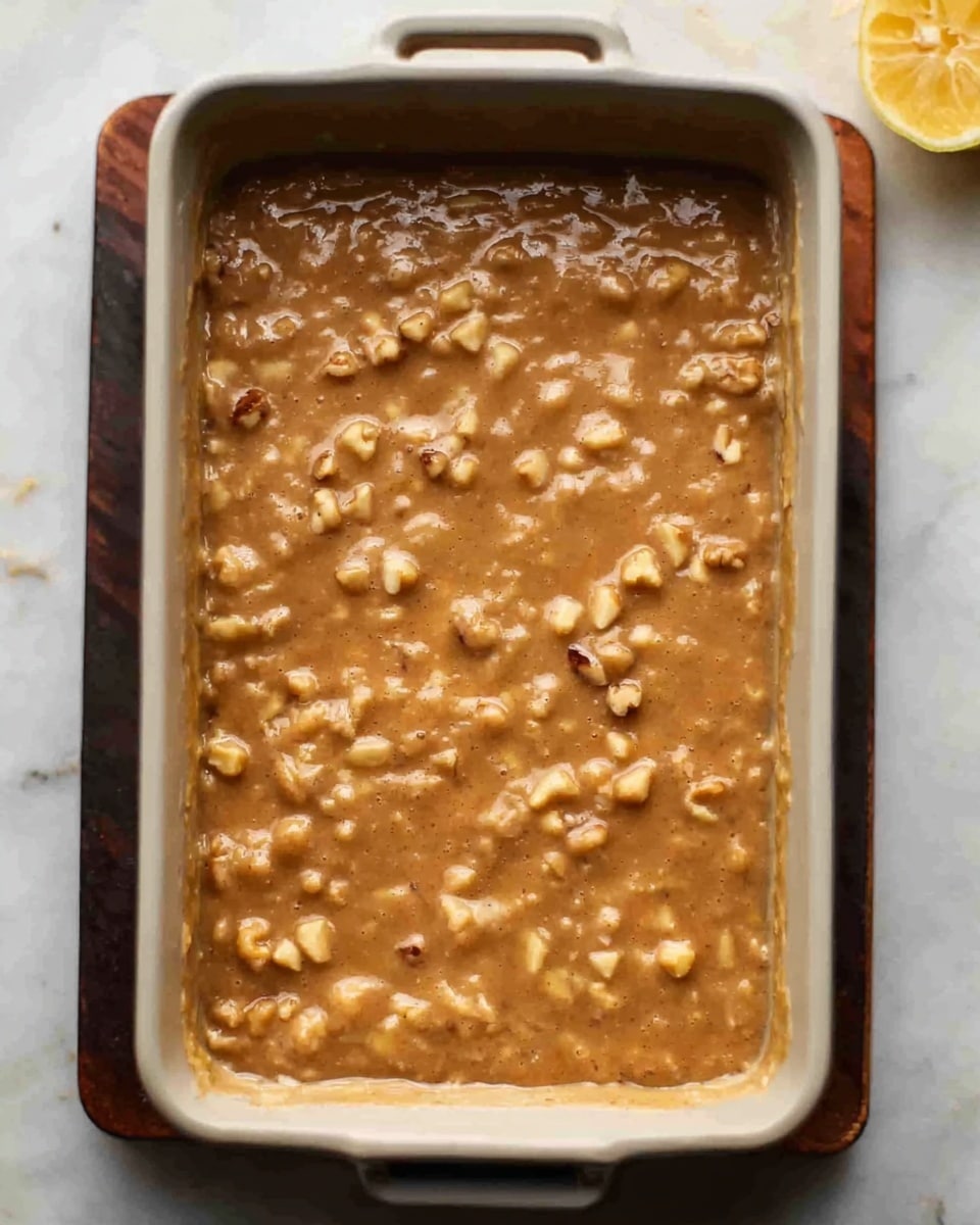 A white rectangular baking dish filled with a thick, light brown batter mixed with visible chunks of nuts scattered throughout. The surface of the batter is uneven, showing a rich texture with small and large nut pieces embedded. The dish is placed on a white marbled surface with a lemon wedge partially visible in the top right corner. photo taken with an iphone --ar 4:5 --v 7