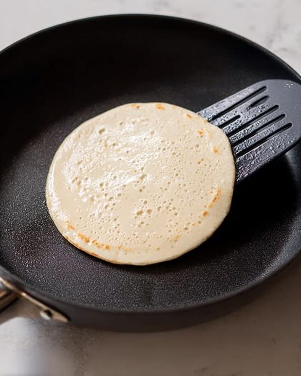 A small, round pancake batter is cooking in a black non-stick pan, with tiny bubbles forming on its surface. The pancake is smooth in texture, mostly light cream color with slight golden spots starting to appear. A black spatula is gently lifting the edge of the pancake from the pan. The background is a white marbled surface with soft lighting highlighting the cooking process. Photo taken with an iphone --ar 4:5 --v 7