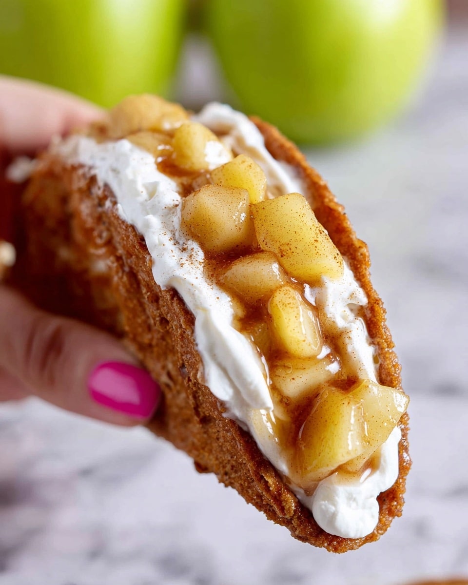 A close-up of a brown taco shell filled with three layers: a bottom layer of white cream, a thick middle layer of small yellow apple pieces covered in shiny golden syrup with bits of cinnamon, and a textured crunchy shell folded into a taco shape held gently by a woman's hand with pink painted nails. The background features a soft white marbled texture with a blurred green apple. photo taken with an iphone --ar 4:5 --v 7