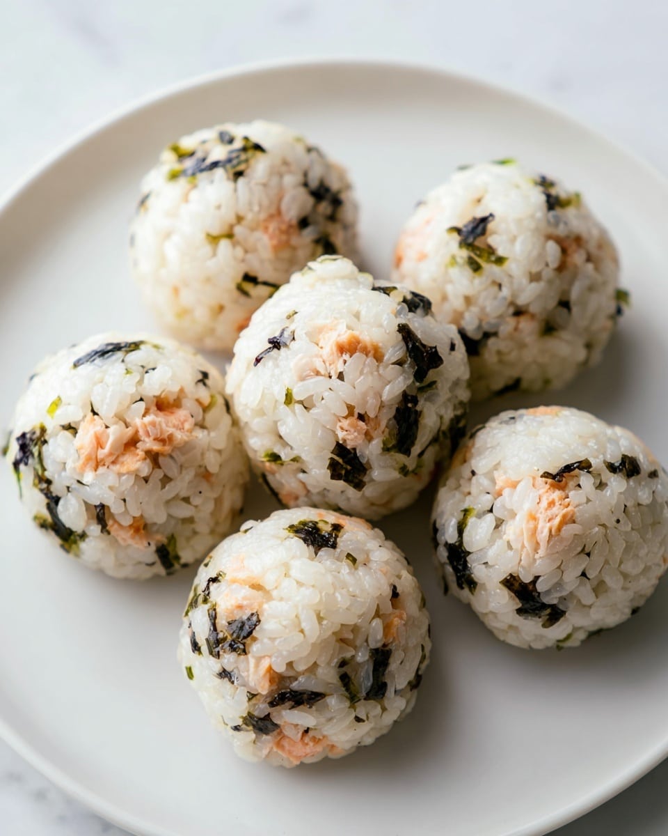 The image shows seven round rice balls arranged on a plain white plate, all placed on a white marbled surface. Each rice ball is made from sticky white rice mixed with small bits of black seaweed and small pink pieces of cooked fish evenly spread throughout. The texture of the rice looks soft and sticky, while the seaweed pieces add a slightly rough and dark contrast, and the fish pieces add a tender, light pink color inside the rice. The rice balls are arranged in a loose cluster with neatly rounded shapes, showing a slight shine from the rice grains. photo taken with an iphone --ar 4:5 --v 7