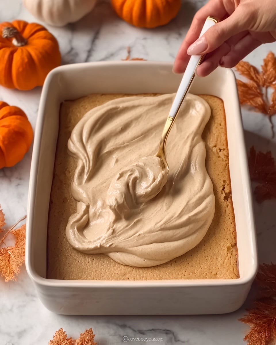 A rectangular white ceramic dish filled with three layers: the bottom layer is a soft beige cake or cookie base, visible around the edges; the middle layer is a slightly darker, orange-brown filling peeking through the edges; the top layer is thick, smooth, and creamy with a light tan color spread evenly across the dish, showing swirled ridges from spreading. A woman's hand holding a white and gold spoon is spreading the creamy top layer in the bottom right corner. The dish is placed on a white marbled surface surrounded by autumn decorations like small orange pumpkins and leaves. Photo taken with an iphone --ar 4:5 --v 7