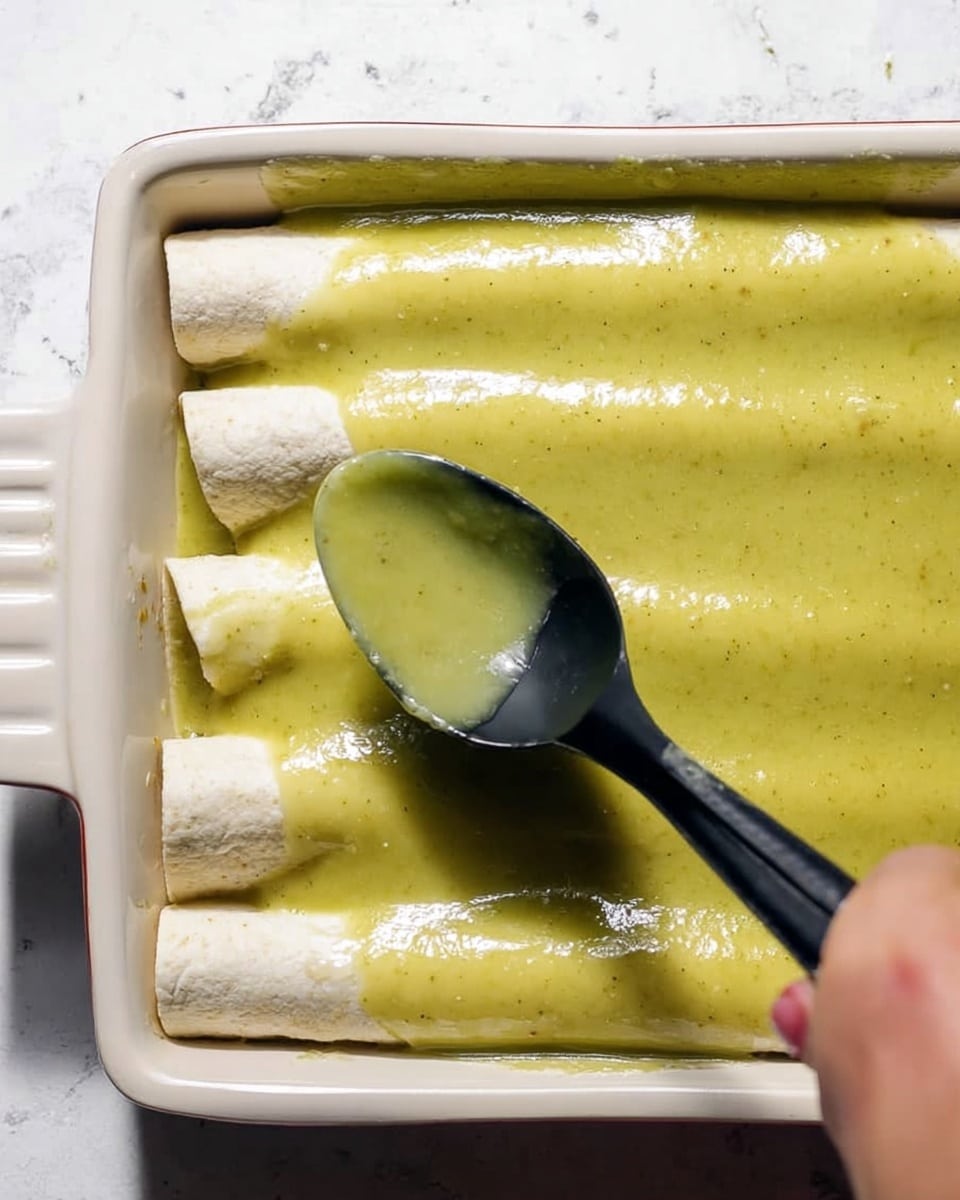 The image shows a close-up of white rolled tortillas arranged side by side inside a white rectangular baking dish. On the top, there is a thick green sauce being spooned over the tortillas with a black spoon, spreading smoothly across the surface. A woman's hand holds the spoon, visible on the right side of the image, gently pouring the sauce. The background beneath the dish is a white marbled texture. The lighting is bright, highlighting the smooth, creamy texture of the sauce and the soft tortillas. photo taken with an iphone --ar 4:5 --v 7