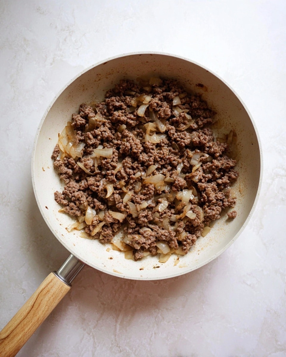 A white pan with a light wood handle holds cooked ground beef mixed with small pieces of cooked onions. The beef is brown and crumbly, and the onions are soft and slightly translucent, all evenly spread across the pan. The pan is placed on a white marbled surface. photo taken with an iphone --ar 4:5 --v 7