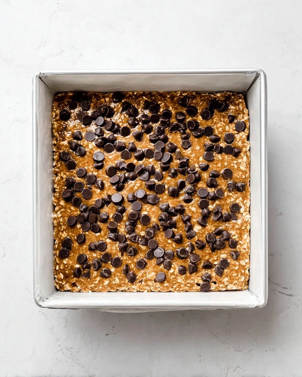 A square white baking pan filled with one layer of light brown oatmeal cookie dough mixed with visible oats, sprinkled evenly on the top with many small, round, dark chocolate chips, sitting on a white marbled surface photo taken with an iphone --ar 4:5 --v 7