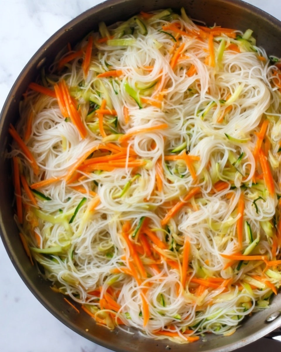 This image shows a pan filled with thin white rice noodles mixed with thin strips of orange carrots and pale green pieces of zucchini. The vegetables and noodles are spread evenly throughout the pan, creating a light and colorful mix with soft textures. The pan sits on a white marbled surface. photo taken with an iphone --ar 4:5 --v 7