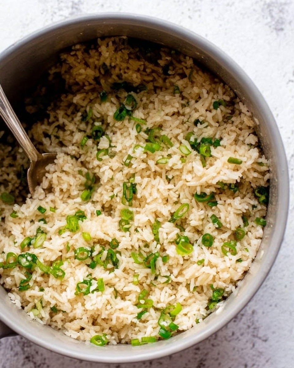 The image shows a close-up of a gray pot filled with cooked rice mixed with small pieces of green scallions. The rice is light brown, fluffy, and evenly mixed with the bright green scallions scattered throughout. A metal spoon is partially visible on the left side inside the pot, suggesting the rice is ready to serve. The background is a white marbled texture. photo taken with an iphone --ar 4:5 --v 7