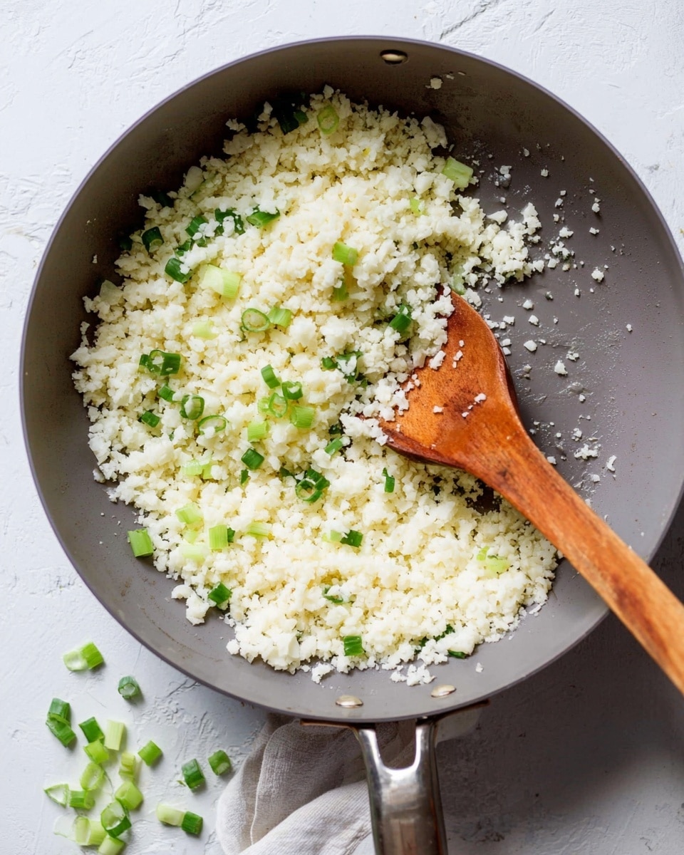 A close-up of a grey pan filled with fluffy cooked cauliflower rice mixed with small pieces of green onion, with a wooden spoon stirring it in the pan. The pan is on a white marbled textured surface, and a few pieces of chopped green onion are scattered around the pan. photo taken with an iphone --ar 4:5 --v 7