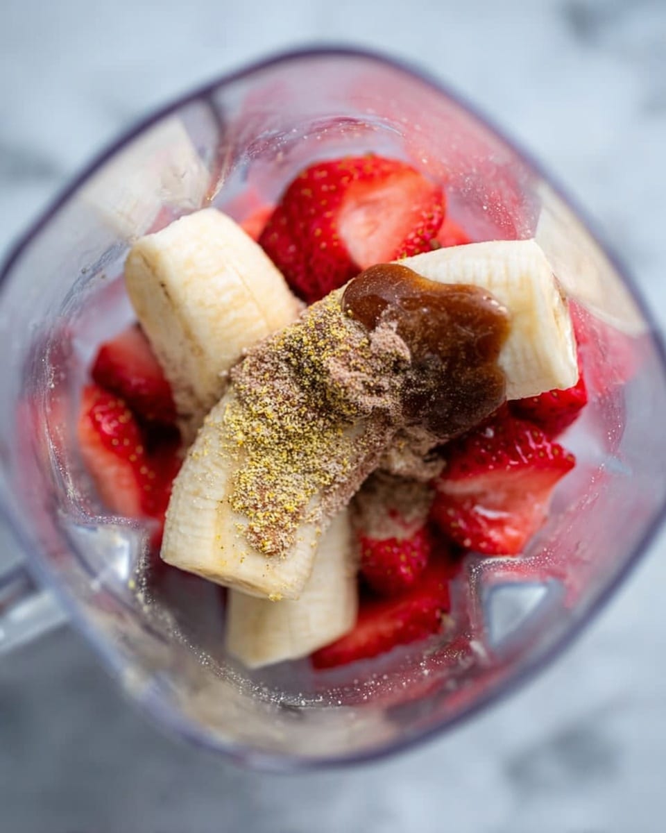 Inside a clear blender jar on a white marbled texture, there are three main layers visible: at the bottom is a pile of sliced bright red strawberries, on top of them lay two peeled banana halves showing light yellow with softer textures, and the topmost layer is a light brown powder sprinkled over the bananas with a glossy dark brown liquid poured partially over the powder and fruit. photo taken with an iphone --ar 4:5 --v 7