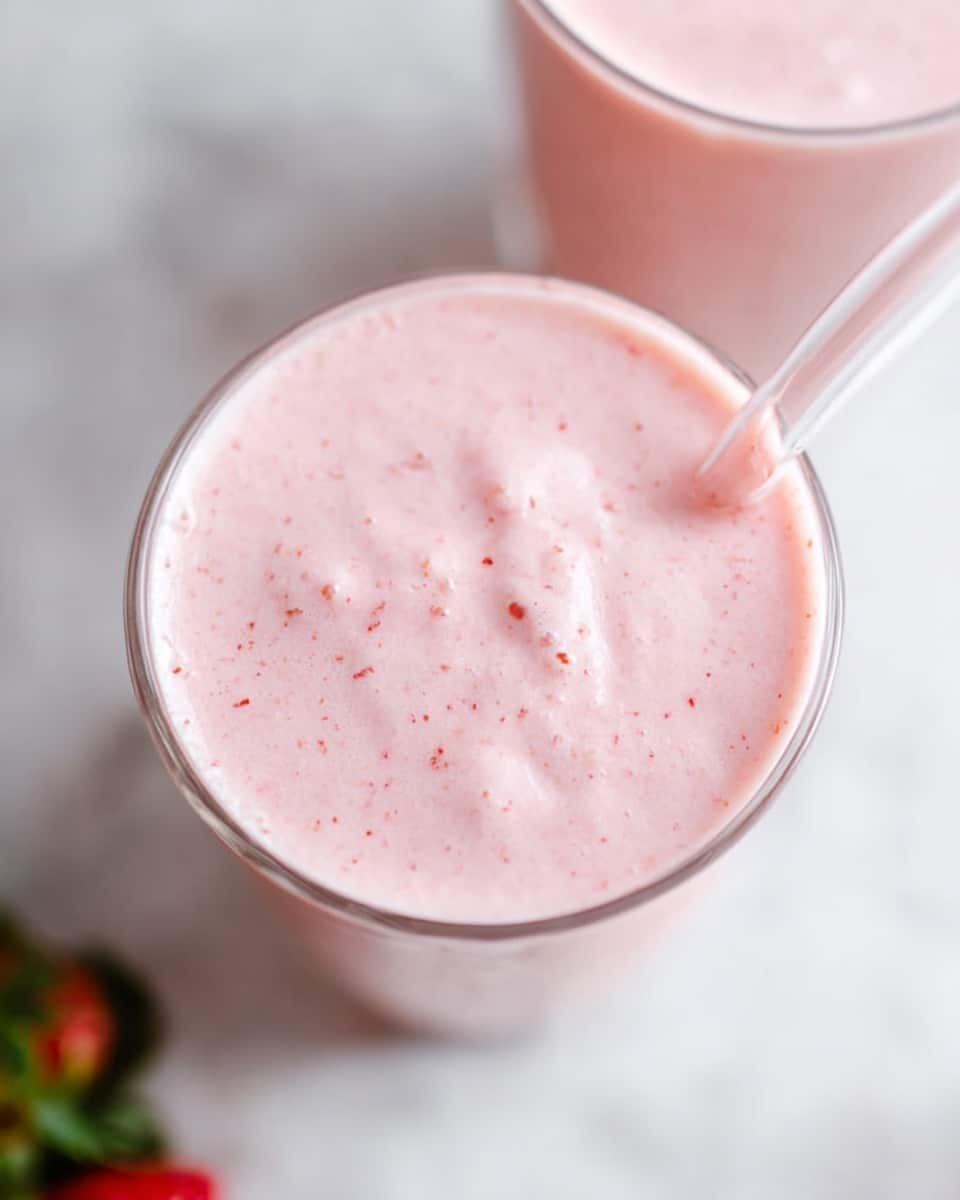 The image shows a close-up top view of a glass filled with a light pink smoothie. The smoothie has a creamy, slightly foamy texture with tiny specks throughout. A clear straw is inserted into the drink, angling slightly to the side. Part of another similar glass is visible in the top right corner. The glasses are placed on a white marbled surface with a small green and red object blurred in the bottom left corner. Photo taken with an iphone --ar 4:5 --v 7