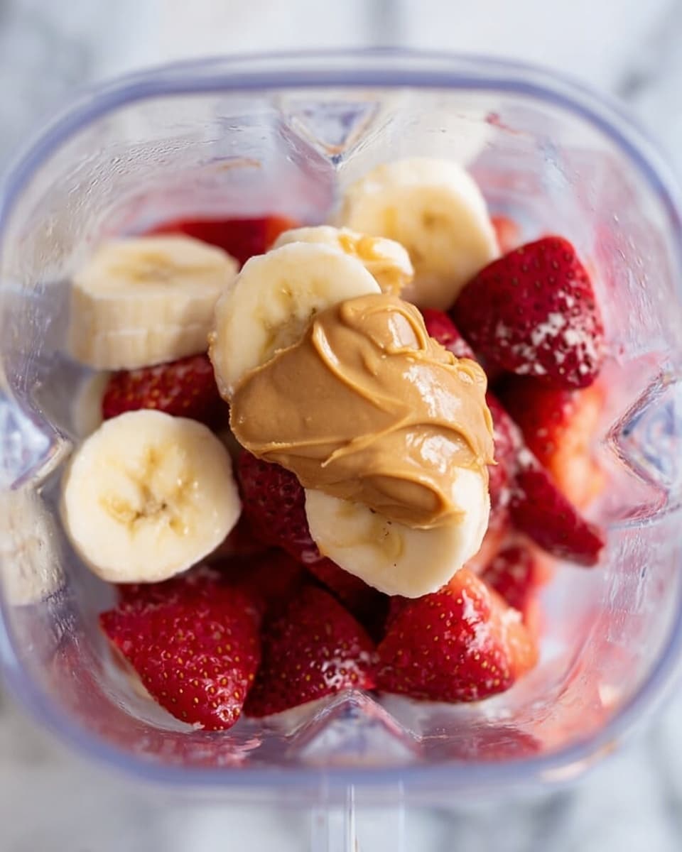 The image shows a clear blender container with several layers inside. The bottom layer is made of chunky red frozen strawberries, followed by round slices of yellow banana that sit on top and slightly around the strawberries. On the top center of the banana slices, there is a thick dollop of light brown peanut butter. The background surface is a white marbled texture. Photo taken with an iphone --ar 4:5 --v 7