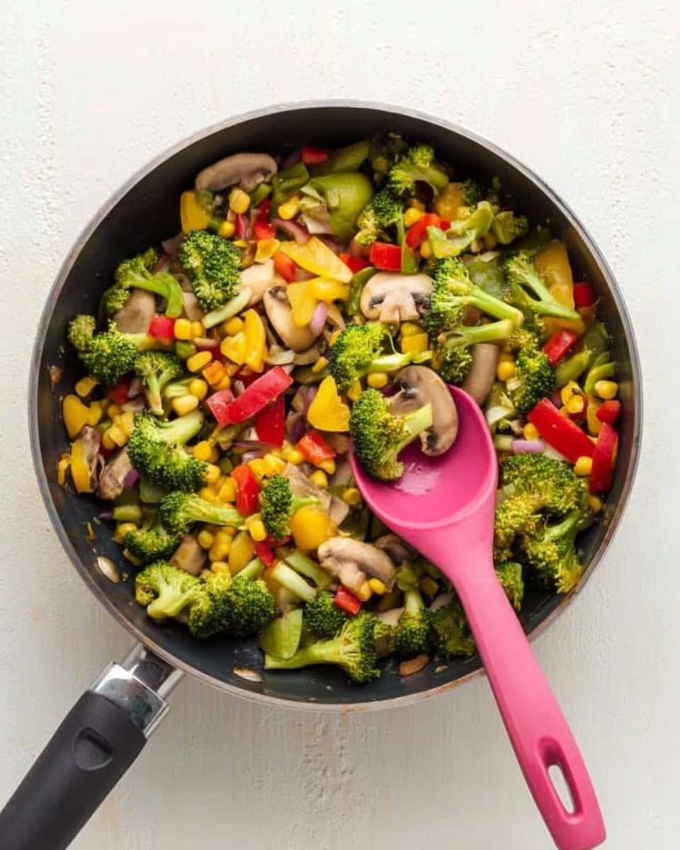 A black frying pan filled with a colorful mix of cooked vegetables including green broccoli, yellow corn, red bell peppers, and light brown mushroom slices. A pink silicone spoon rests inside the pan, angled over the vegetables. The pan sits on a white marbled surface, creating a clean and bright background. The vegetables have a fresh, slightly glossy look from being cooked. Photo taken with an iphone --ar 4:5 --v 7