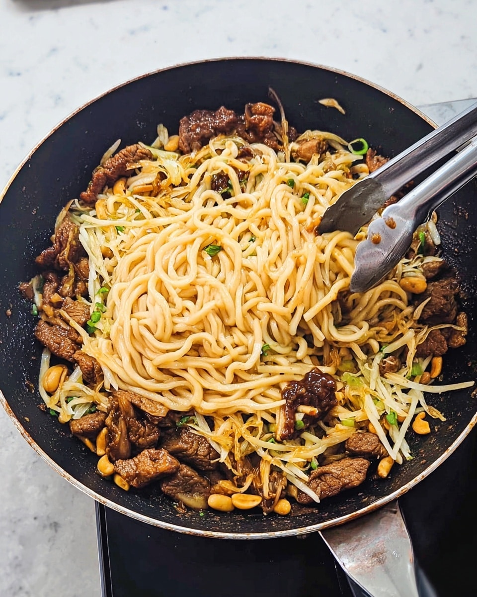 A black frying pan contains a mix of thick beige noodles, some coated with a dark brown sauce sitting at the center, surrounded by golden brown cooked pieces of meat scattered around the edges. Small yellow peanuts and thin white shredded cheese are sprinkled throughout, with tiny green bits adding a touch of color. A metal spaghetti serving tool is positioned in the pan, holding a cluster of noodles. The surface beneath the pan is a white marbled texture. Photo taken with an iphone --ar 4:5 --v 7
