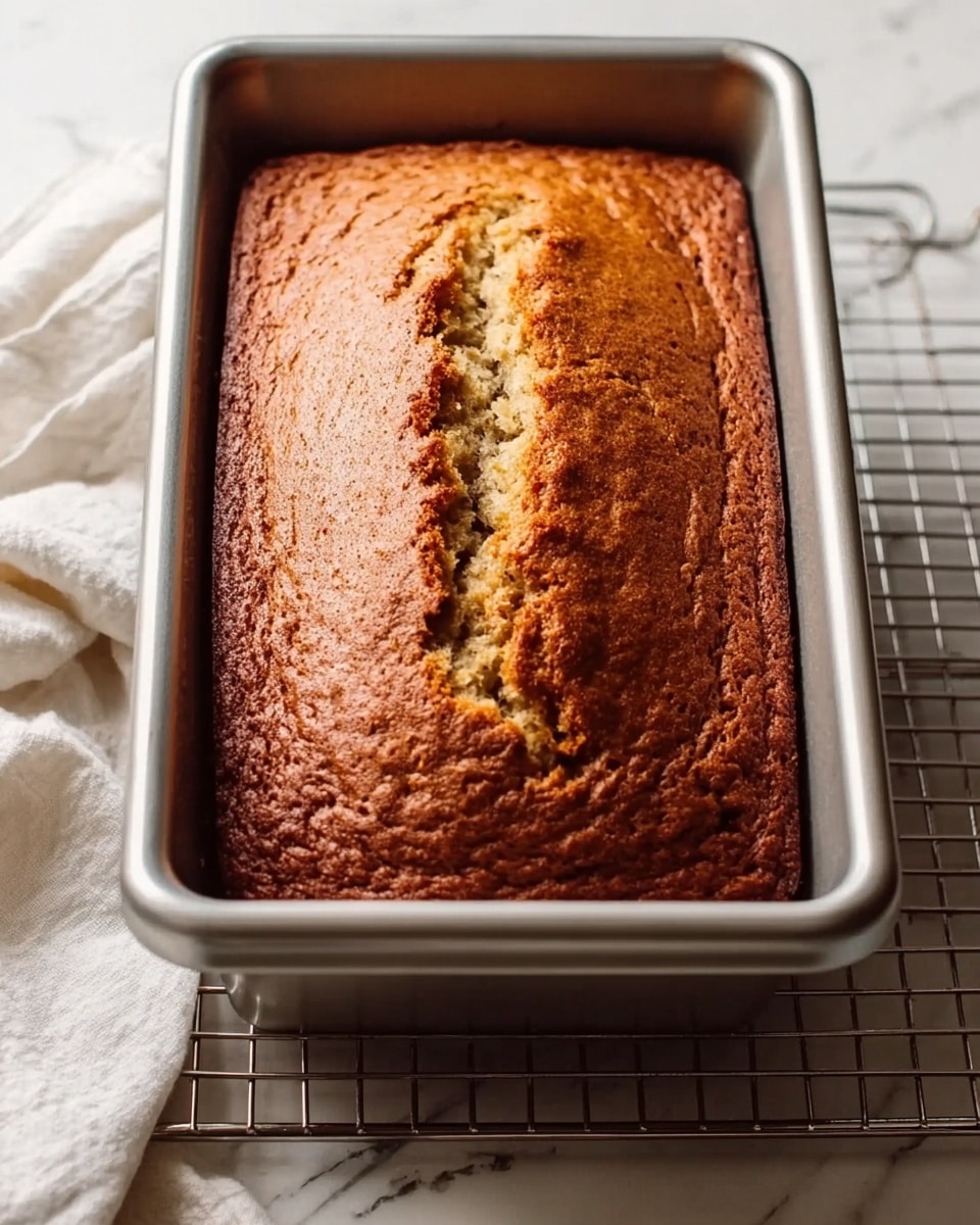 A freshly baked loaf of banana bread sits in a silver metal baking pan. The top of the bread is golden brown with a deep crack running down the center, showing a slightly darker, textured crust. The pan rests on a wire cooling rack placed over a white marbled surface, and a white cloth is casually spread next to it. The lighting highlights the soft, moist texture of the bread, with subtle shadows adding depth. photo taken with an iphone --ar 4:5 --v 7