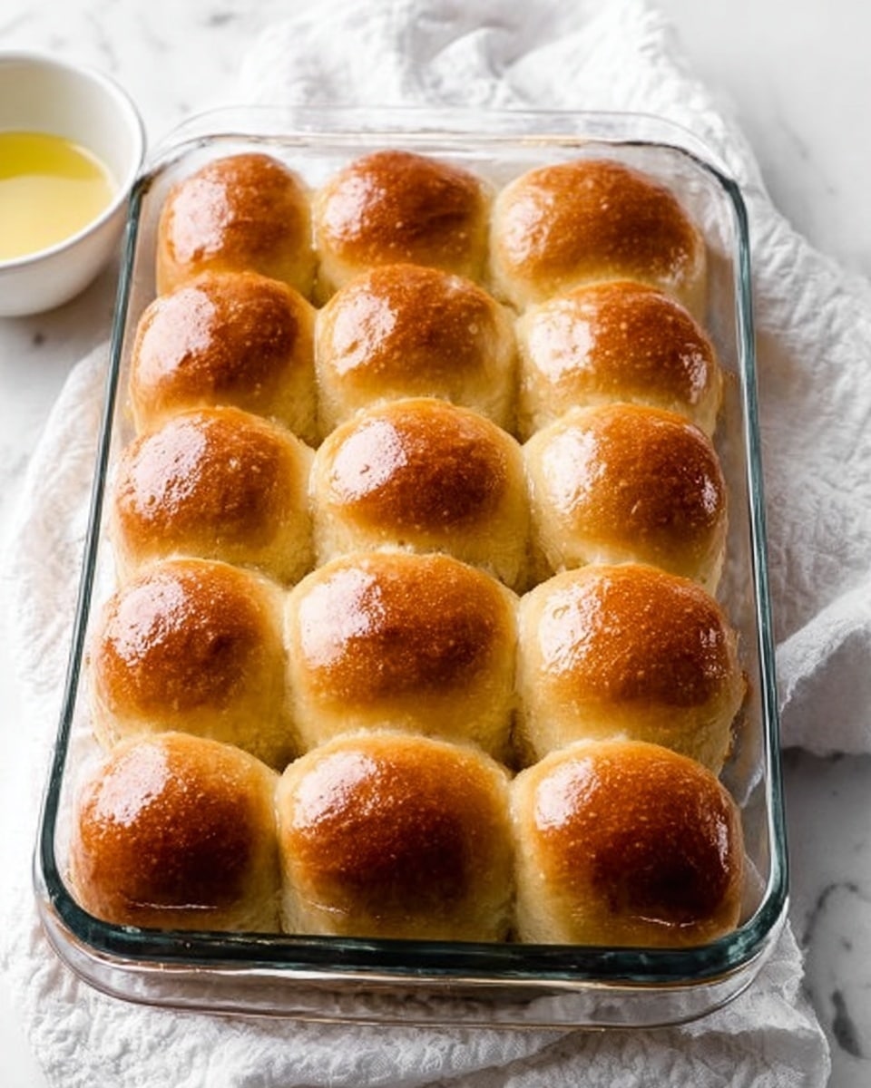 A clear glass baking dish holds fifteen golden brown dinner rolls arranged in a 3 by 5 grid. Each roll is soft and shiny with a smooth, slightly glossy top, showing a fresh and fluffy texture under the golden crust. The dish sits on a white marbled surface with a white cloth underneath, adding softness to the scene. To the left side, there is a partial view of a small white bowl that contains a light yellow liquid, possibly butter. The lighting is bright and natural, highlighting the warmth and inviting look of the rolls. photo taken with an iphone --ar 4:5 --v 7
