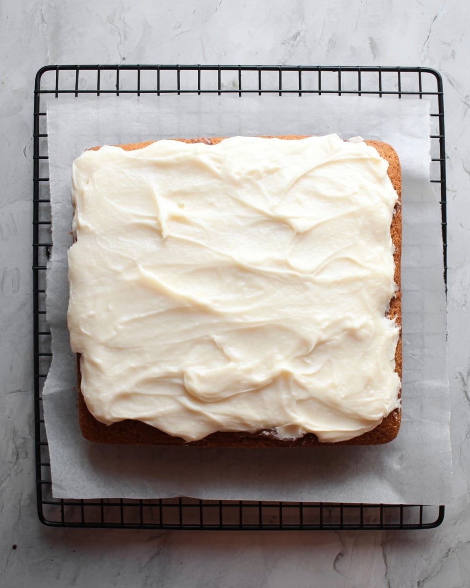 A square cake with one visible layer is placed on a white marbled surface with parchment paper underneath and a cooling rack beneath it. The cake layer is light brown and topped with a thick, uneven layer of white cream spread across the top, showing smooth and slightly textured swirls. The edges of the cream layer extend close to the cake's sides but do not fully cover them. Photo taken with an iphone --ar 4:5 --v 7