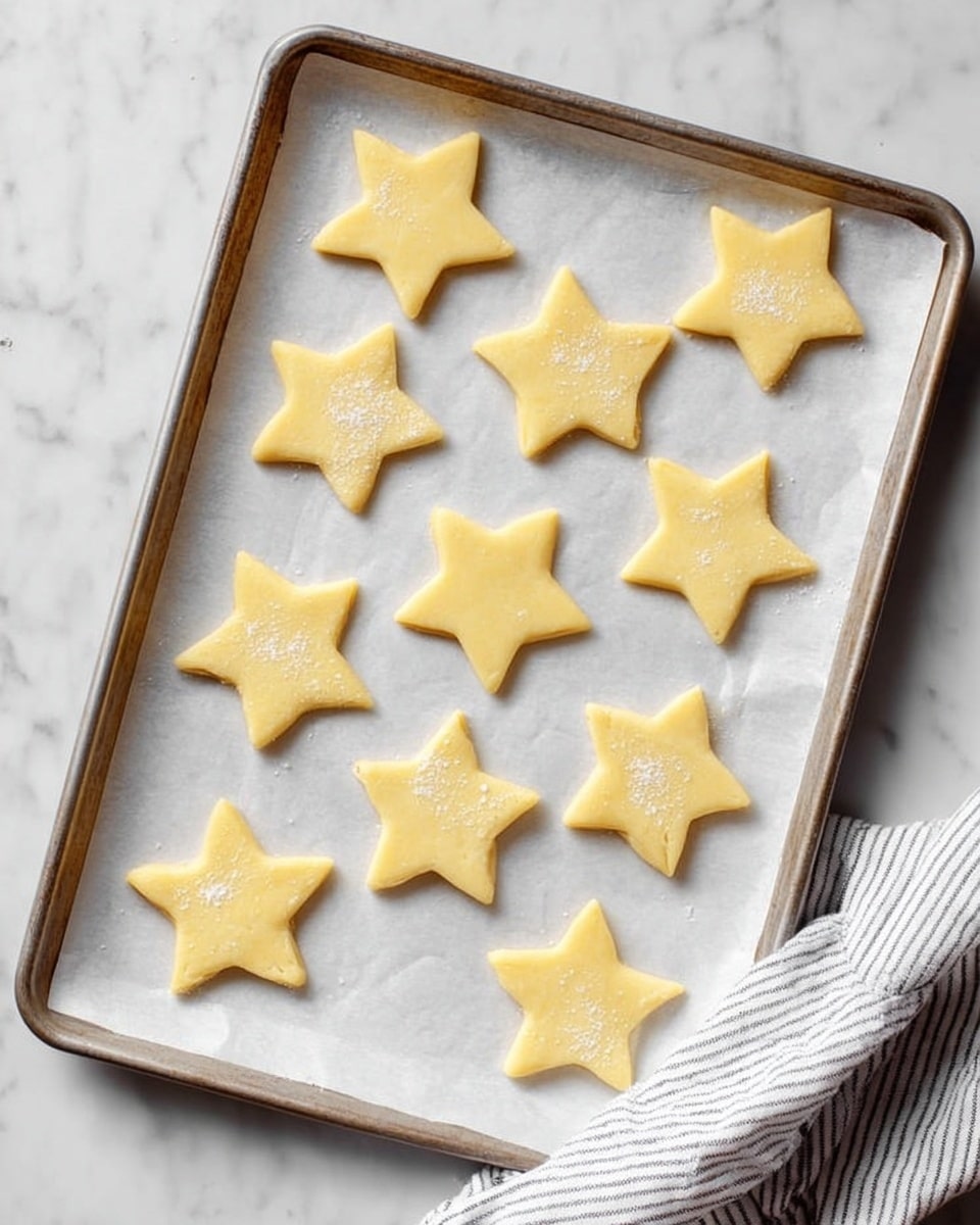 The image shows a baking tray lined with white parchment paper, placed on a white marbled surface. On the parchment paper, there are thirteen star-shaped dough pieces evenly spread out. The dough is pale yellow with a smooth texture, and each star is neatly cut with five points. Some stars have a slight dusting of flour or sugar on top, adding a light texture contrast. A white and gray striped cloth is seen partially at the bottom right corner. Photo taken with an iphone --ar 4:5 --v 7