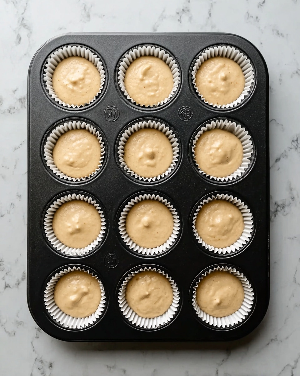 A black metal muffin tray holds twelve white paper liners filled with light beige batter. Each liner is filled nearly to the top with smooth batter that has small air bubbles and soft peaks visible on the surface. The tray rests on a white marbled surface with subtle gray veins, showing a clean and bright background. The batter looks ready to bake, filling each muffin cup evenly without spilling. photo taken with an iphone --ar 4:5 --v 7