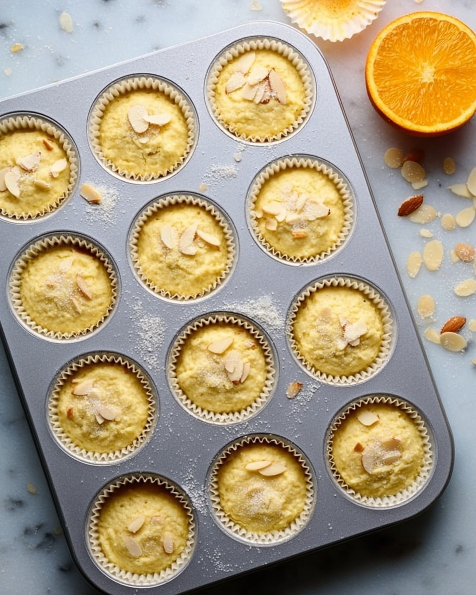 A silver muffin tray holding twelve paper-lined white cupcake molds filled with light yellow, rough batter. Some cupcakes have thin, off-white almond slices scattered on top, while others are bare. A shiny, coarse sugar topping is lightly sprinkled across the cupcakes and tray. To the right, part of a bright orange half is visible on a white marbled surface, with a few almond slices and sugar grains scattered around the tray. photo taken with an iphone --ar 4:5 --v 7