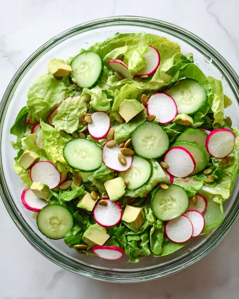 A clear glass bowl filled with fresh green lettuce leaves as the base layer, topped with evenly spread slices of green cucumber and thin, round slices of radish with white centers and red edges. Scattered small cubes of pale green avocado sit on top, along with a few light brown pumpkin seeds sprinkled across for texture. The bowl is placed on a white marbled surface, showing a colorful and fresh salad mix without any dressing. photo taken with an iphone --ar 4:5 --v 7