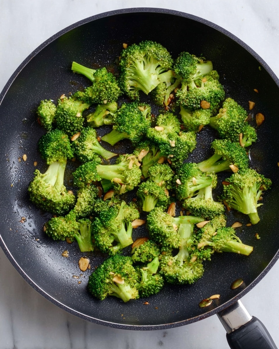 The image shows a black frying pan filled with small, bright green broccoli pieces that are slightly browned on the edges. Scattered among the broccoli are thin, light brown slices of garlic that add texture. The pan itself has a slightly rough surface with a few drops of oil visible, and it sits on a white marbled surface. The broccoli pieces are spread out evenly in one layer inside the pan. photo taken with an iphone --ar 4:5 --v 7
