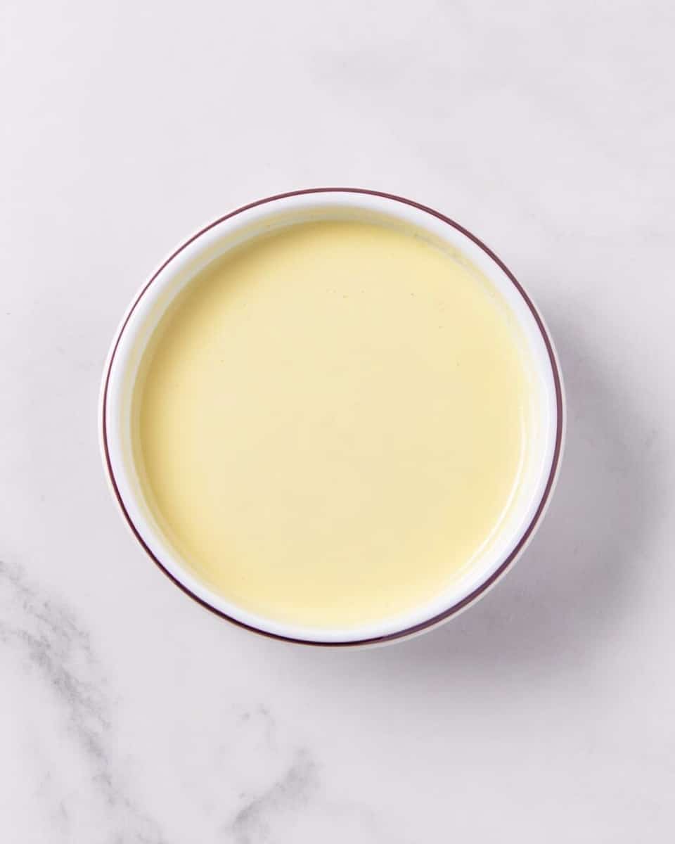 A round white bowl filled with a smooth, pale yellow creamy liquid, evenly spread without any visible texture or bubbles, placed on a white marbled surface. photo taken with an iphone --ar 4:5 --v 7