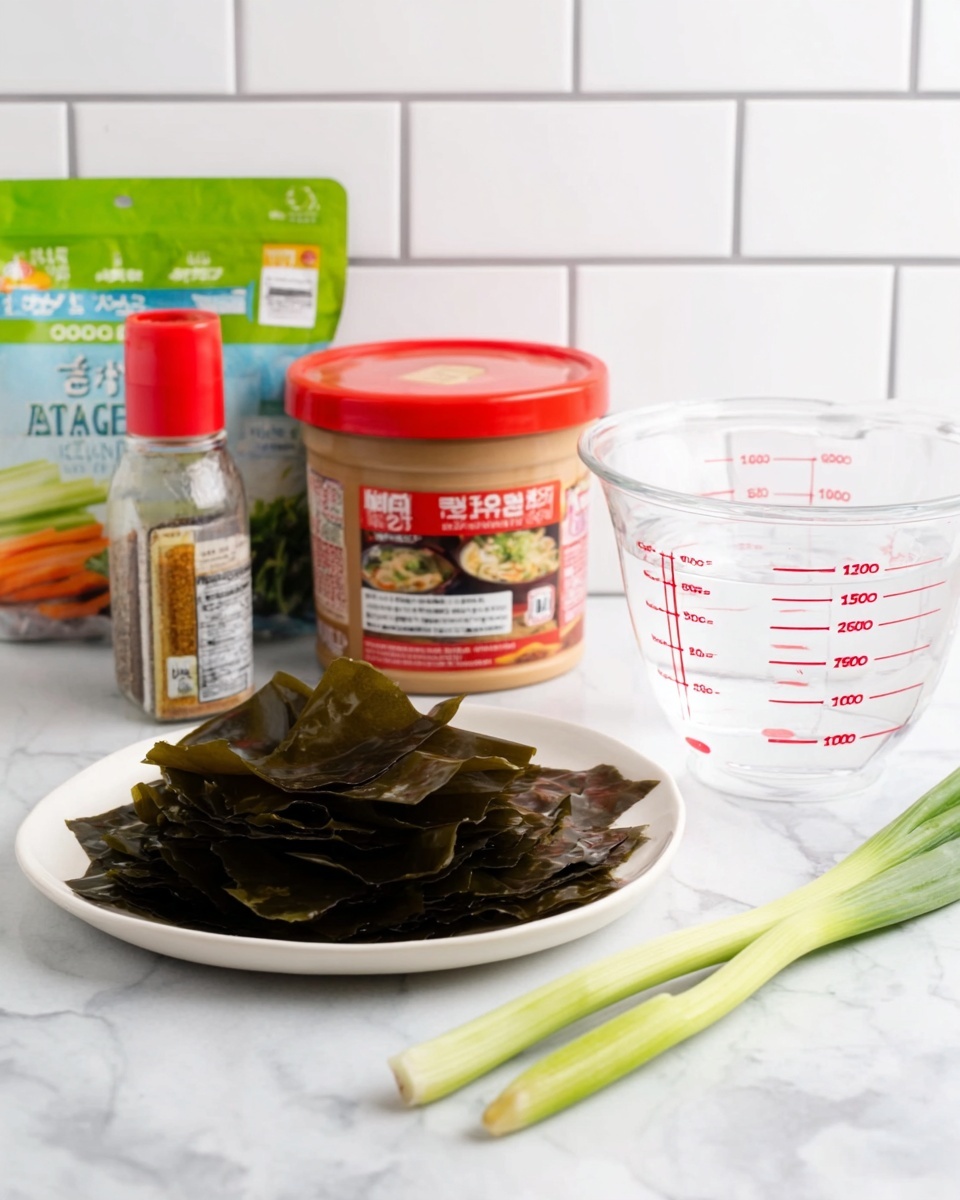 The image shows a white plate filled with dark green dried seaweed sheets stacked loosely. Behind the plate, there is a container of soybean paste with a red label and lid, showing an image of soup on the front. Next to the soybean paste, there is a spice bottle with a red cap, and behind that is a clear measuring cup with red measurement marks filled partially with water. A fresh green onion lies flat on a white marbled surface, close to the front of the plate. In the background, to the left, there is a package with a photo of sliced vegetables and a green top section, all set against white subway tiles. The photo taken with an iphone --ar 4:5 --v 7