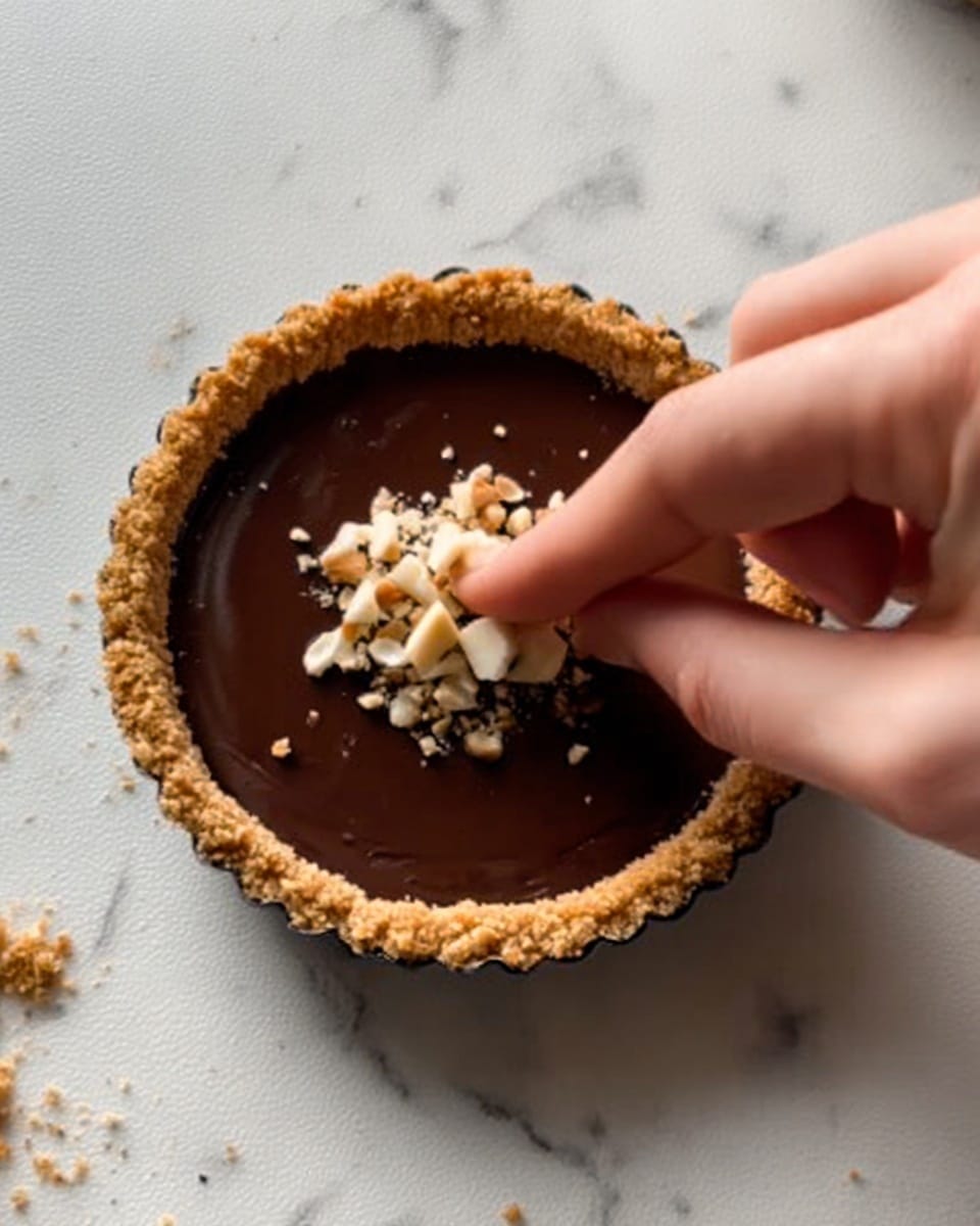 A small tart with a golden crumbly crust filled with smooth dark chocolate ganache. On top, a woman's hand is placing small pieces of white and light brown crushed nuts in the center. The tart sits on a white marbled surface with subtle gray veins, and the texture of the tart crust looks rough and crunchy. Photo taken with an iphone --ar 4:5 --v 7
