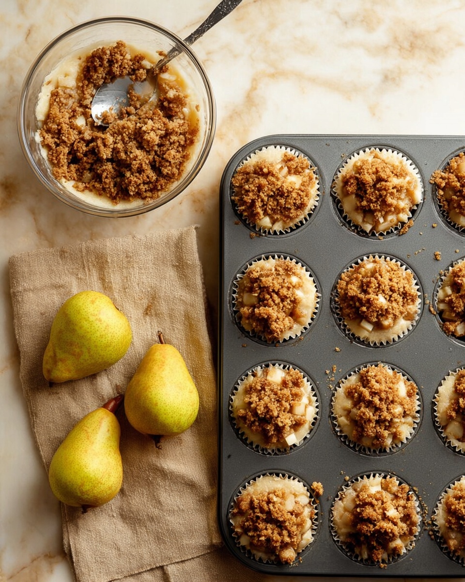 A metal muffin tray holds eight cups filled with a two-layer dessert: the bottom layer is a light, creamy mixture dotted with pear pieces, topped with a golden brown crumbly streusel layer that has a rough, crunchy texture. To the left of the tray, a clear bowl contains more of the crumbly topping with a silver spoon resting inside on a beige cloth. Beside the cloth, two ripe pears with yellow-green skin lie on a white marbled surface. The colors are warm and inviting, with the crumb layer showing different shades of brown and the pears adding natural green and yellow tones. photo taken with an iphone --ar 4:5 --v 7
