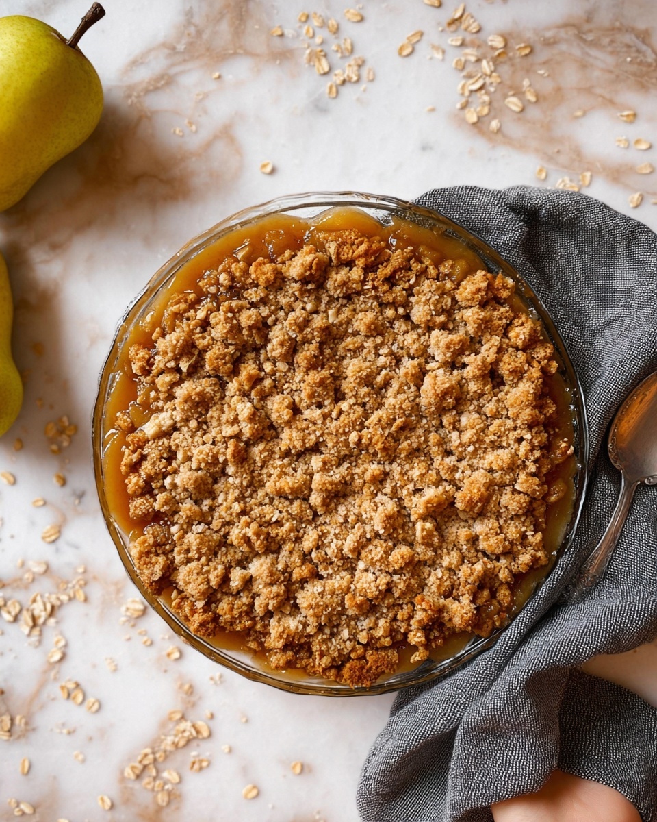 The image shows a round glass dish filled with a two-layer dessert. The bottom layer is a soft fruit filling with a shiny, sticky texture in a warm amber color that slightly spills over the edge. The top layer is a thick crumbly topping made up of golden brown oats and small, rough clusters of baked dough, giving it a crunchy look. The dish sits on a white marbled surface with scattered oat flakes around it. Nearby, there is a gray cloth, a silver spoon, and a woman’s hand holding the dish gently from one side. A yellow pear is partially visible on the left side of the image. Photo taken with an iphone --ar 4:5 --v 7