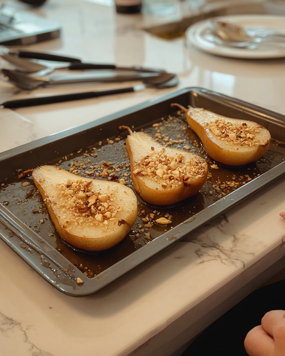 On a black baking tray resting on a white marbled surface, there are four pear halves arranged in two rows with the fleshy side facing up. Each pear half has a golden brown skin with a soft, creamy off-white inside. The pear halves are topped with small pieces of light beige chopped nuts and a shiny layer of golden syrup or honey, which glistens on the surface. Some chopped nuts are scattered around the tray, adding texture. The scene shows part of a woman's dark sleeve and silver utensils on the right side, with clear glass bowls visible in the upper left background. Photo taken with an iphone --ar 4:5 --v 7
