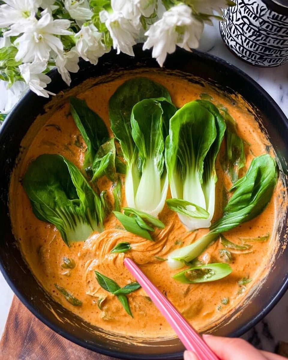 A black bowl holds a thick, creamy orange sauce with a smooth texture filling most of the bowl. Inside the sauce, several fresh green bok choy leaves and white stems are placed on top and partially submerged. A woman's hand with a pink spoon is stirring the sauce, creating a swirl effect near the center of the bowl. The bowl is set on a white marbled surface with white flowers and a black-and-white patterned container in the background. photo taken with an iphone --ar 4:5 --v 7